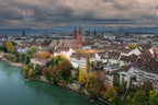 A City Held, aerial photograph of Basel, Switzerland, showing the Basler Münster, historic rooftops and autumn trees beside the Rhine beneath dramatic clouds.