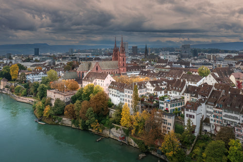 A City Held, aerial photograph of Basel, Switzerland, showing the Basler Münster, historic rooftops and autumn trees beside the Rhine beneath dramatic clouds.