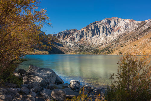 Long-exposure view of Convict Lake, with smooth turquoise water, a rocky shore, autumn leaves, and rugged mountains beneath a clear blue sky.