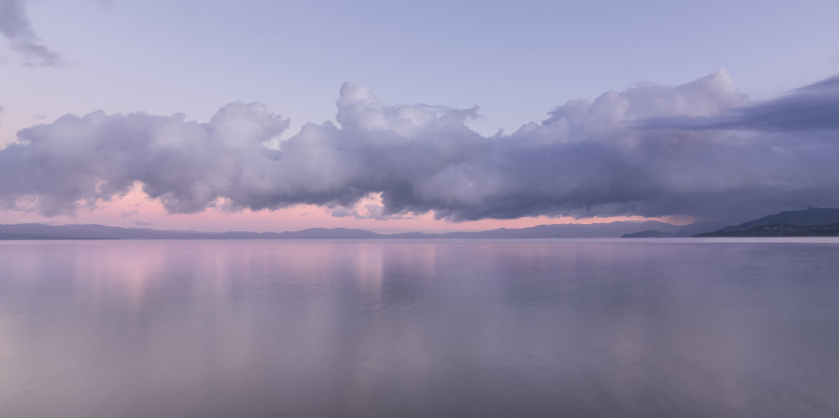 A wide view across Lake Tahoe at sunrise after snowfall, with a dramatic bank of clouds above the distant shoreline and soft pink light reflected in the calm water.