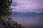 A rocky, pine-framed Lake Tahoe shoreline beneath a lavender sky, with snow-dusted mountains across still water after a morning snowfall.