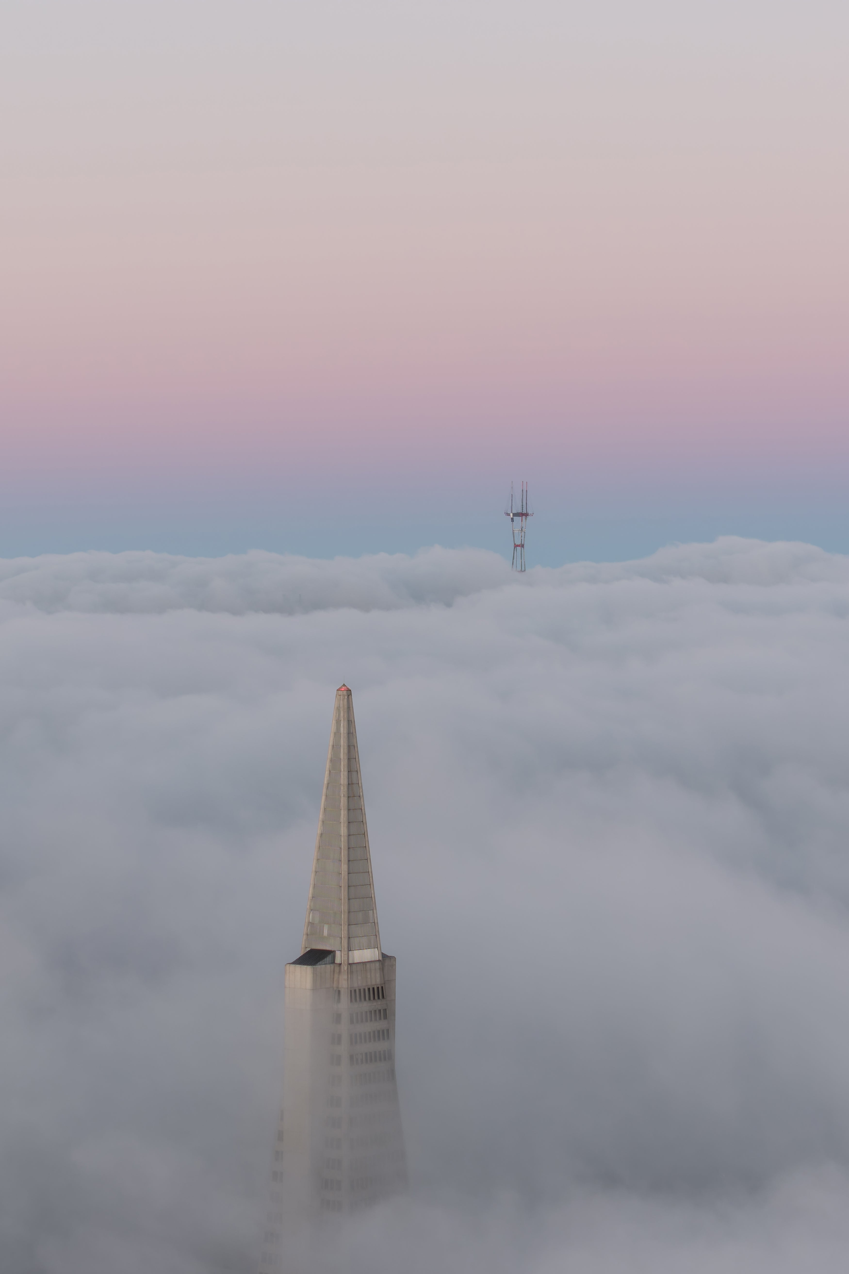 Aerial view of San Francisco’s Transamerica Pyramid and Sutro Tower emerging through layered fog beneath a pastel pink and blue sky.