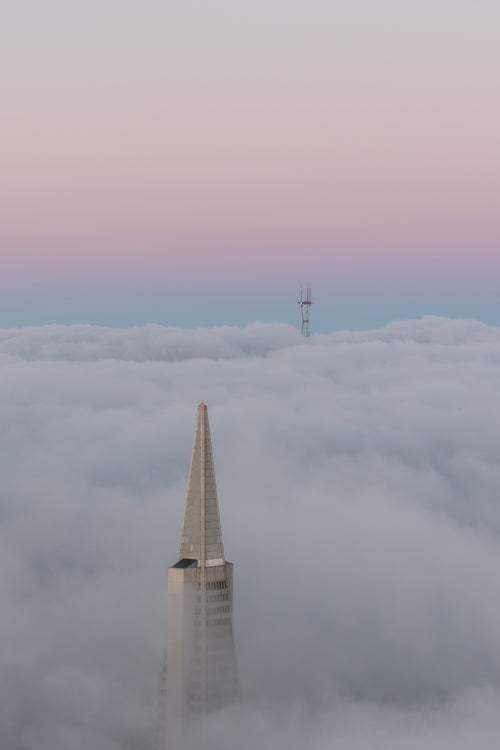 Aerial view of San Francisco’s Transamerica Pyramid and Sutro Tower emerging through layered fog beneath a pastel pink and blue sky.