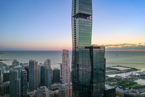 Aerial view above the Chicago skyline at sunrise, with a glass skyscraper extending beyond the frame over Lake Michigan and Navy Pier.