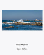 Held Motion open-edition coastal photograph of a wave suspended above rugged rocks amid layered blue sea and sky.
