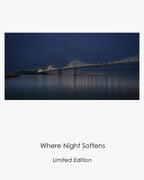 San Francisco–Oakland Bay Bridge illuminated before dawn, with lights and subtle colors reflected across calm water beneath a fog-softened blue sky.