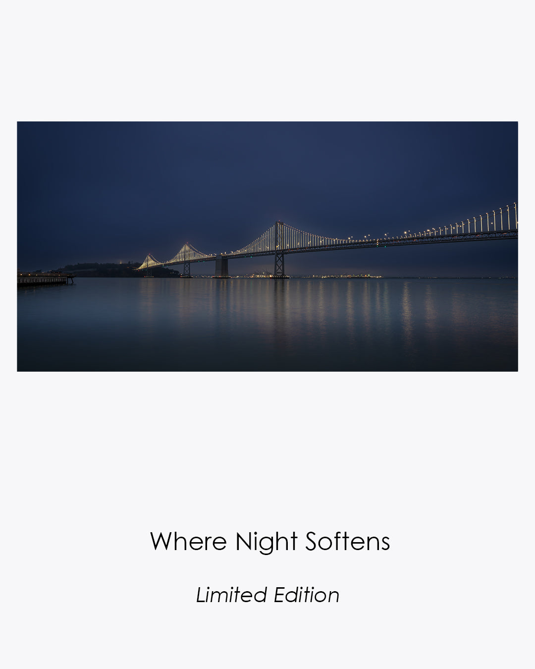 San Francisco–Oakland Bay Bridge illuminated before dawn, with lights and subtle colors reflected across calm water beneath a fog-softened blue sky.