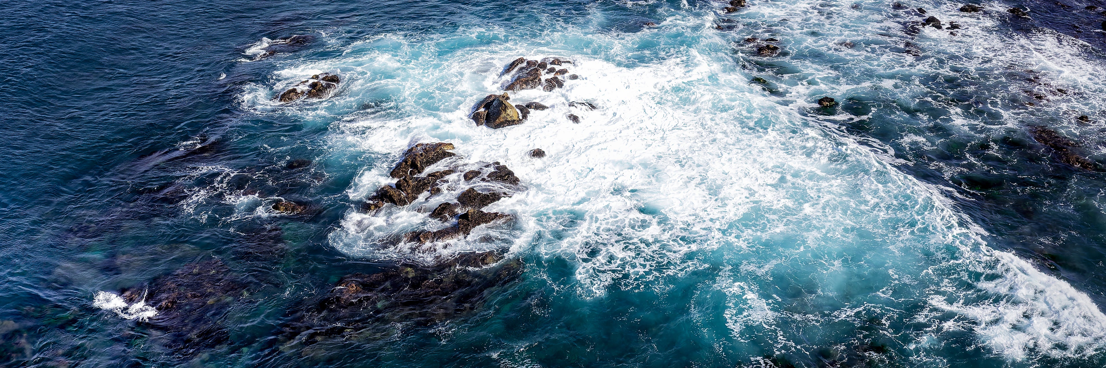 Panoramic aerial photograph of ocean currents swirling around rocks as deep navy water shifts into teal, turquoise, and white foam.