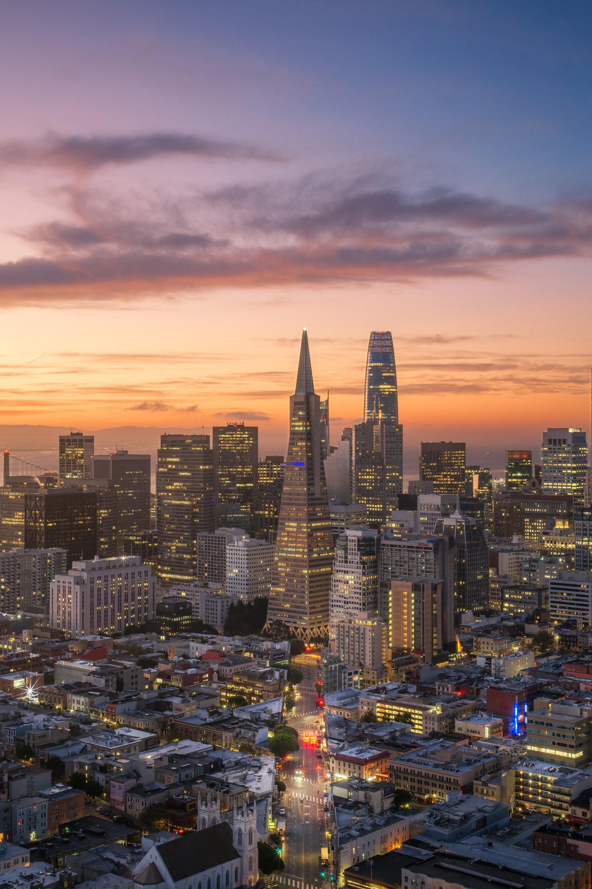 San Francisco at sunrise from above, with Columbus Avenue leading from Saints Peter and Paul Church toward the Transamerica Pyramid and Salesforce Tower beneath a pink-blue sky.