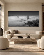 Black-and-white panoramic aerial photograph of both Golden Gate Bridge towers rising through billowing fog above San Francisco Bay.