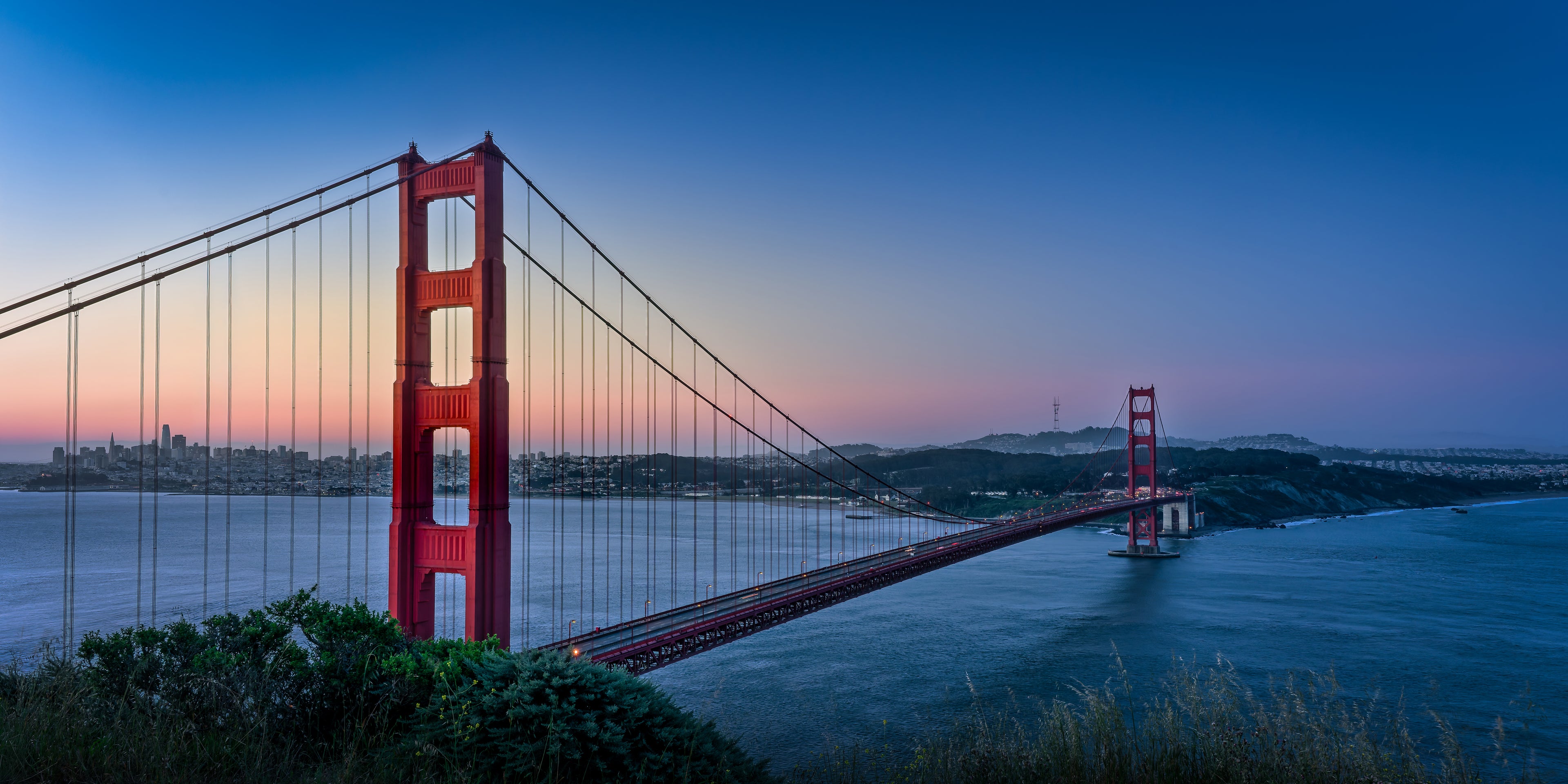 The Golden Gate Bridge glowing against a blue and rose-colored dawn sky with the San Francisco skyline beyond.