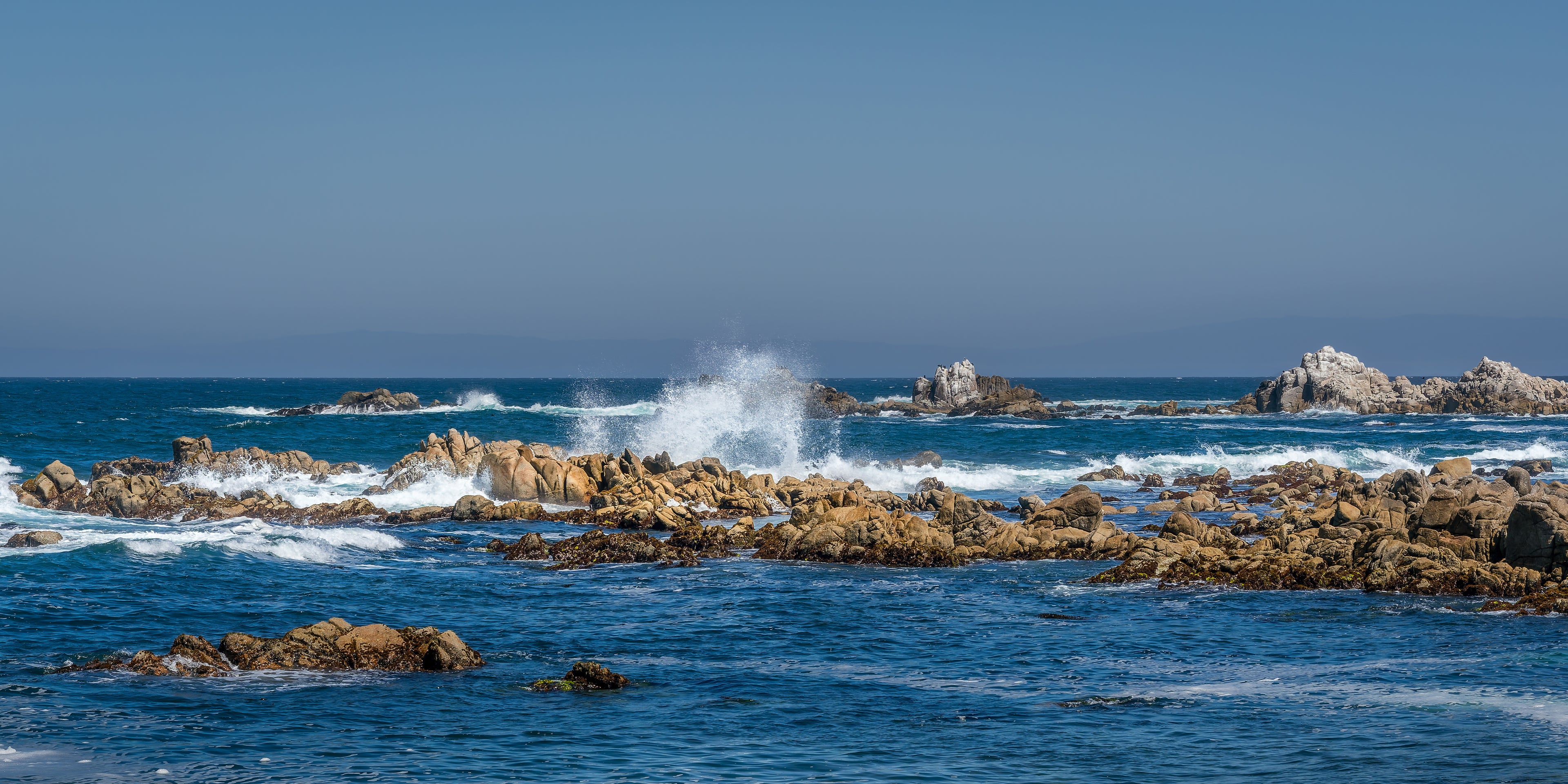 Panoramic coastal photograph of a breaking wave suspended above warm rock formations beneath a softly blended blue sky.