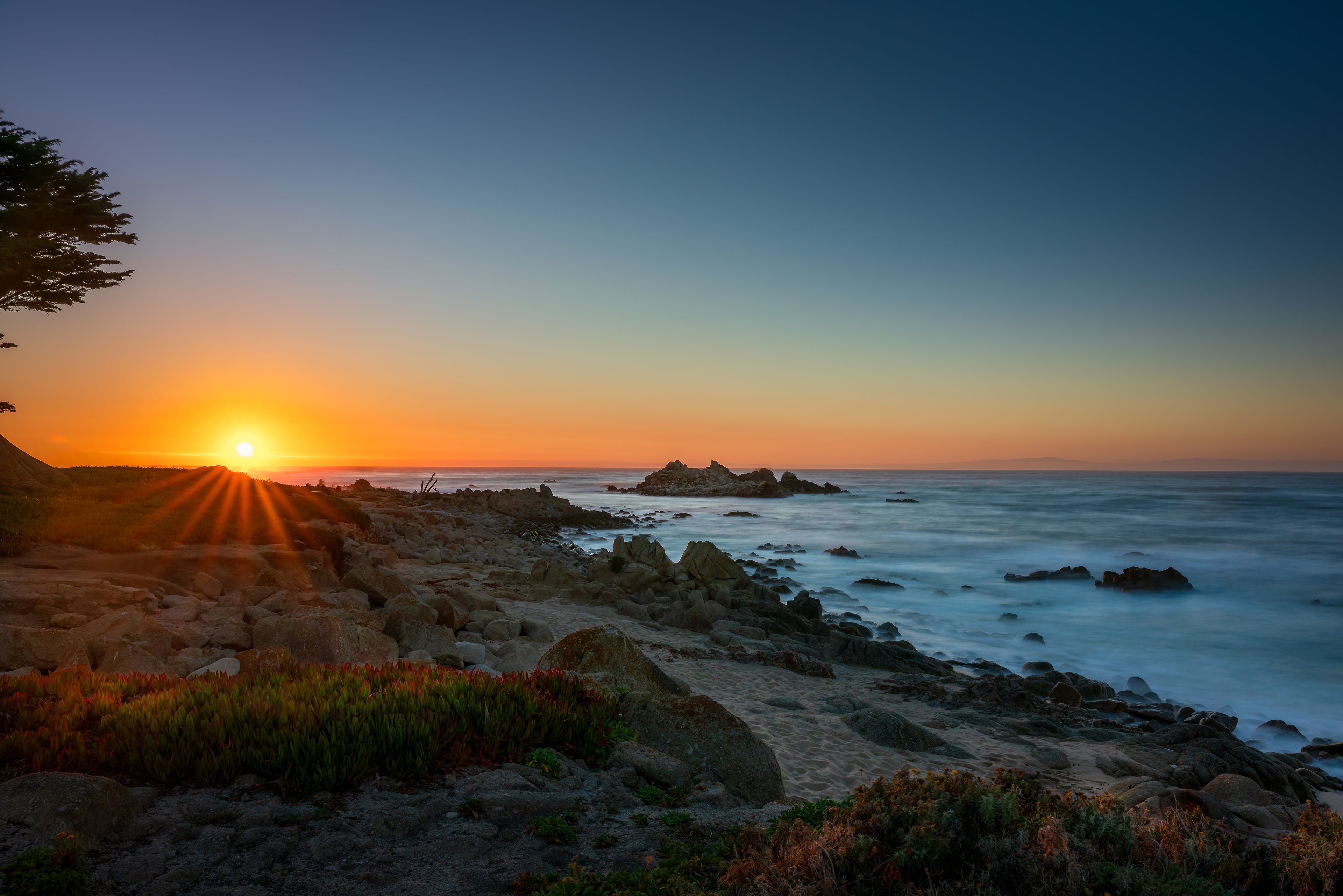 The final sun touching the horizon, casting golden rays across coastal rocks and softly flowing blue water.