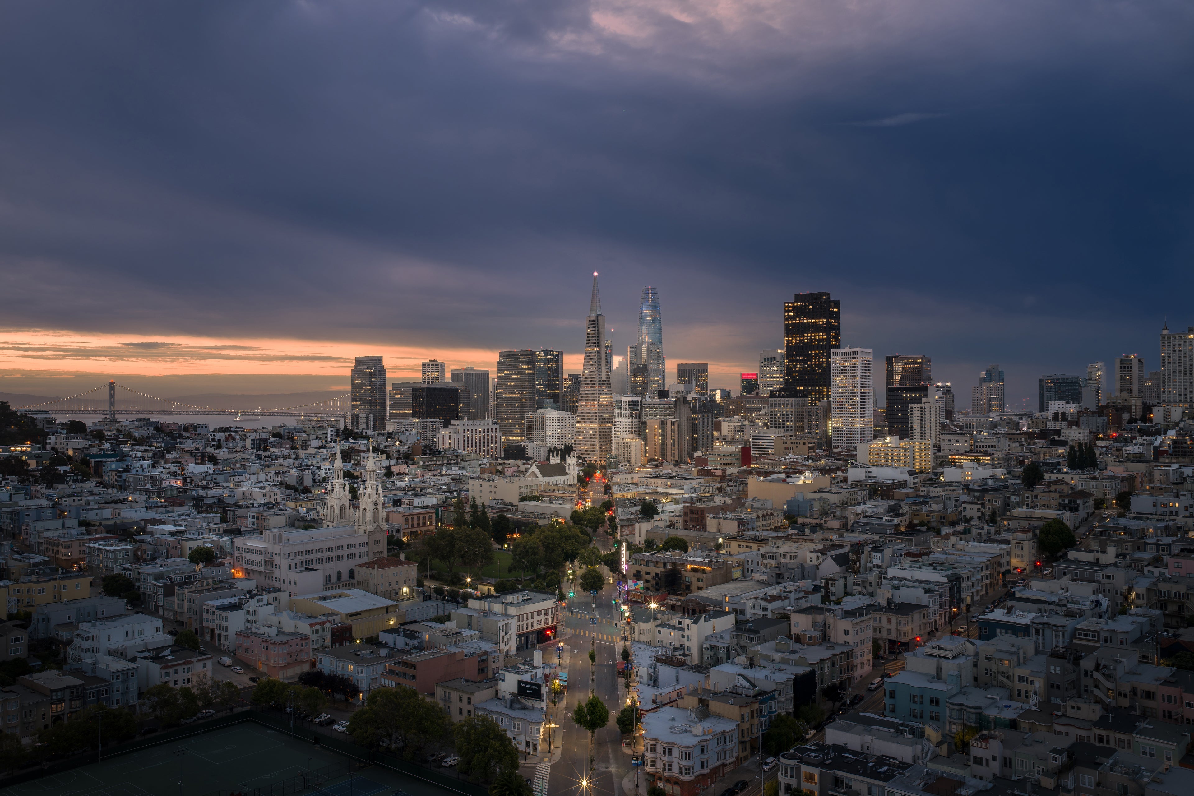 Panoramic San Francisco cityscape at dawn, with the lights of Columbus Avenue drawing the eye toward the Transamerica Pyramid beneath dramatic blue clouds.