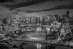 Black-and-white aerial view of an empty Oracle Park glowing beneath dramatic clouds and the San Francisco skyline.