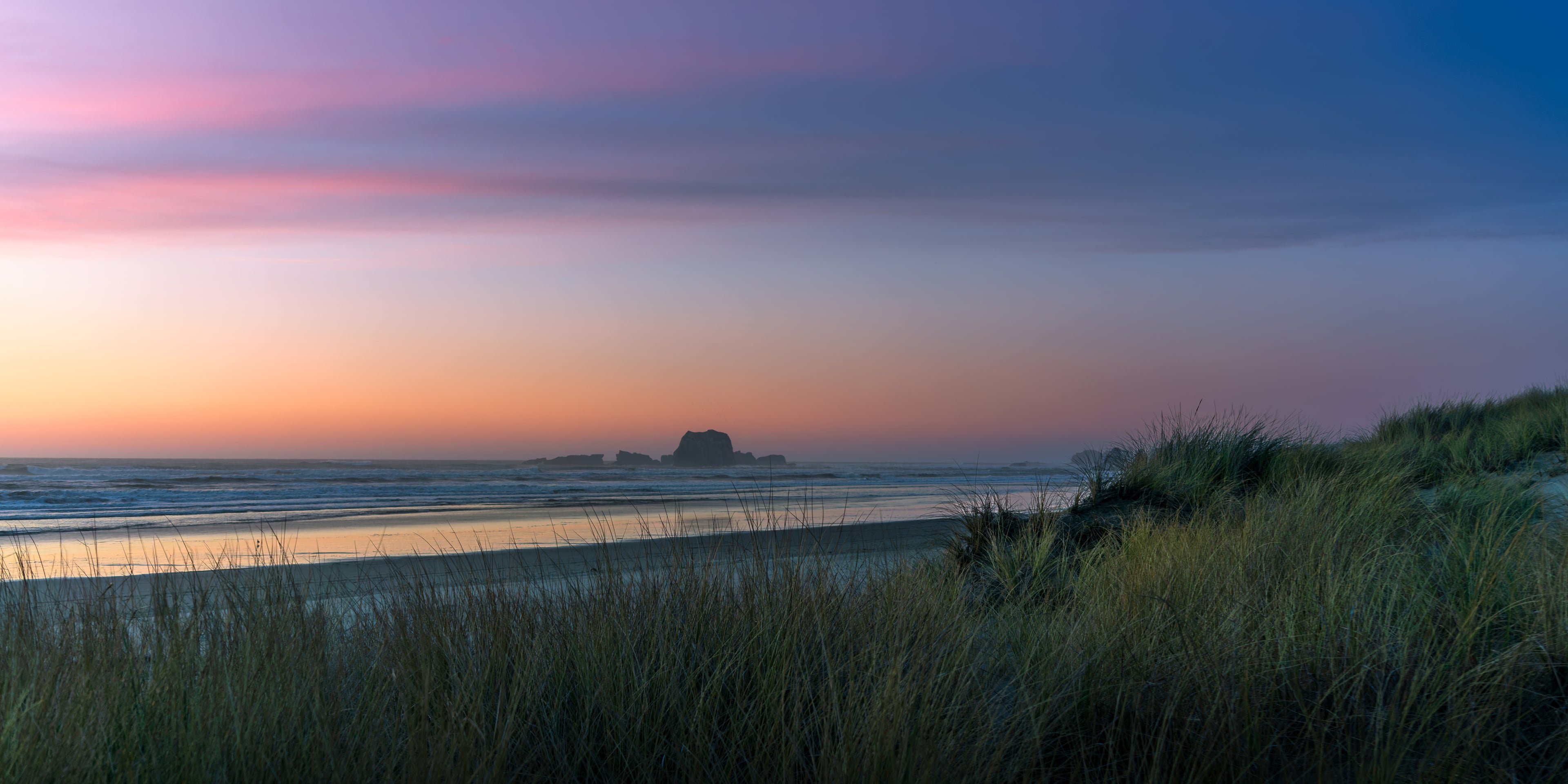 Soft pink and lavender twilight above Oregon coastal sea stacks, reflected water, and windswept dune grass.