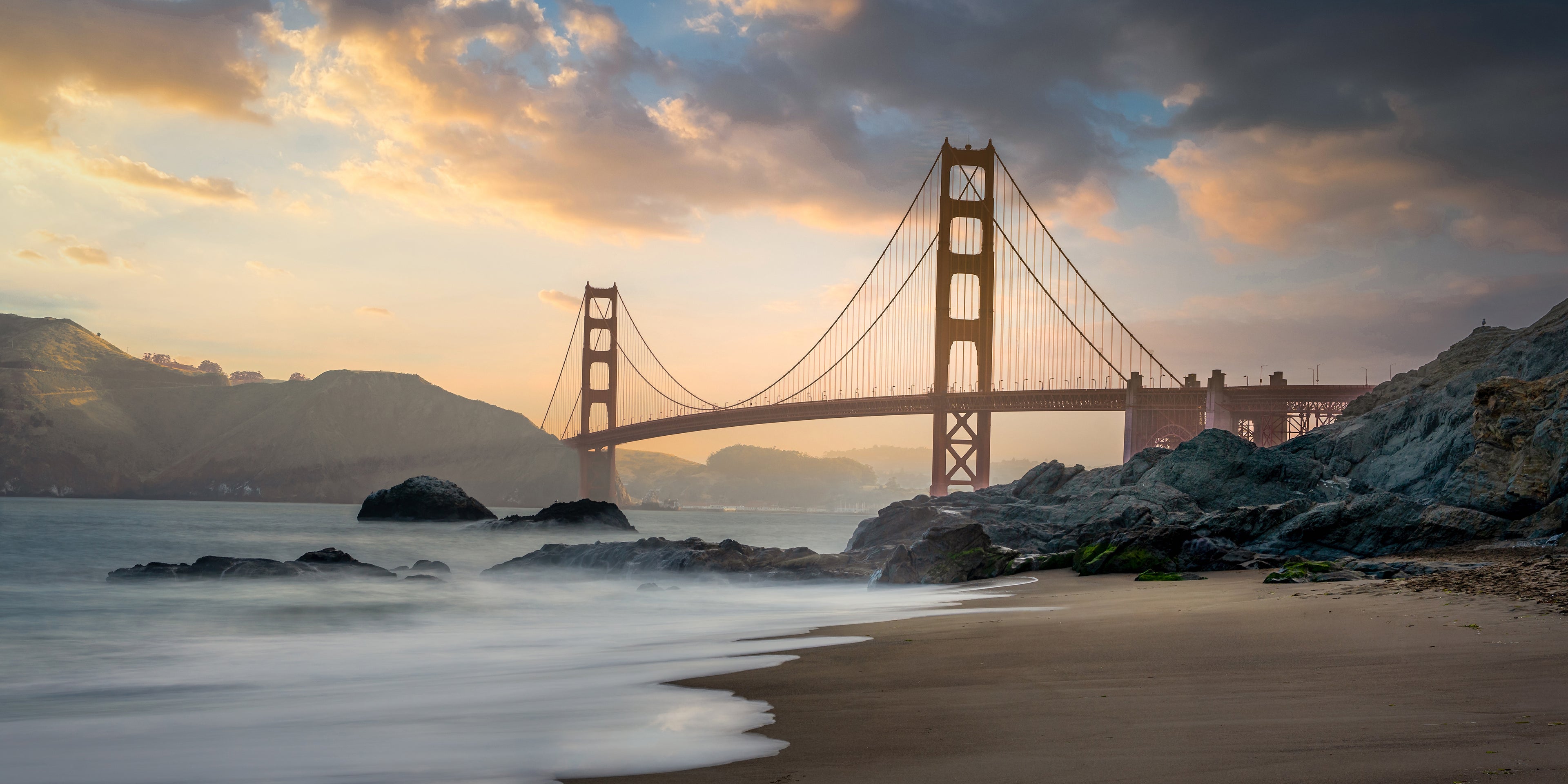 Golden morning light illuminating the Golden Gate Bridge as soft waves reach the rocky shoreline of Baker Beach.