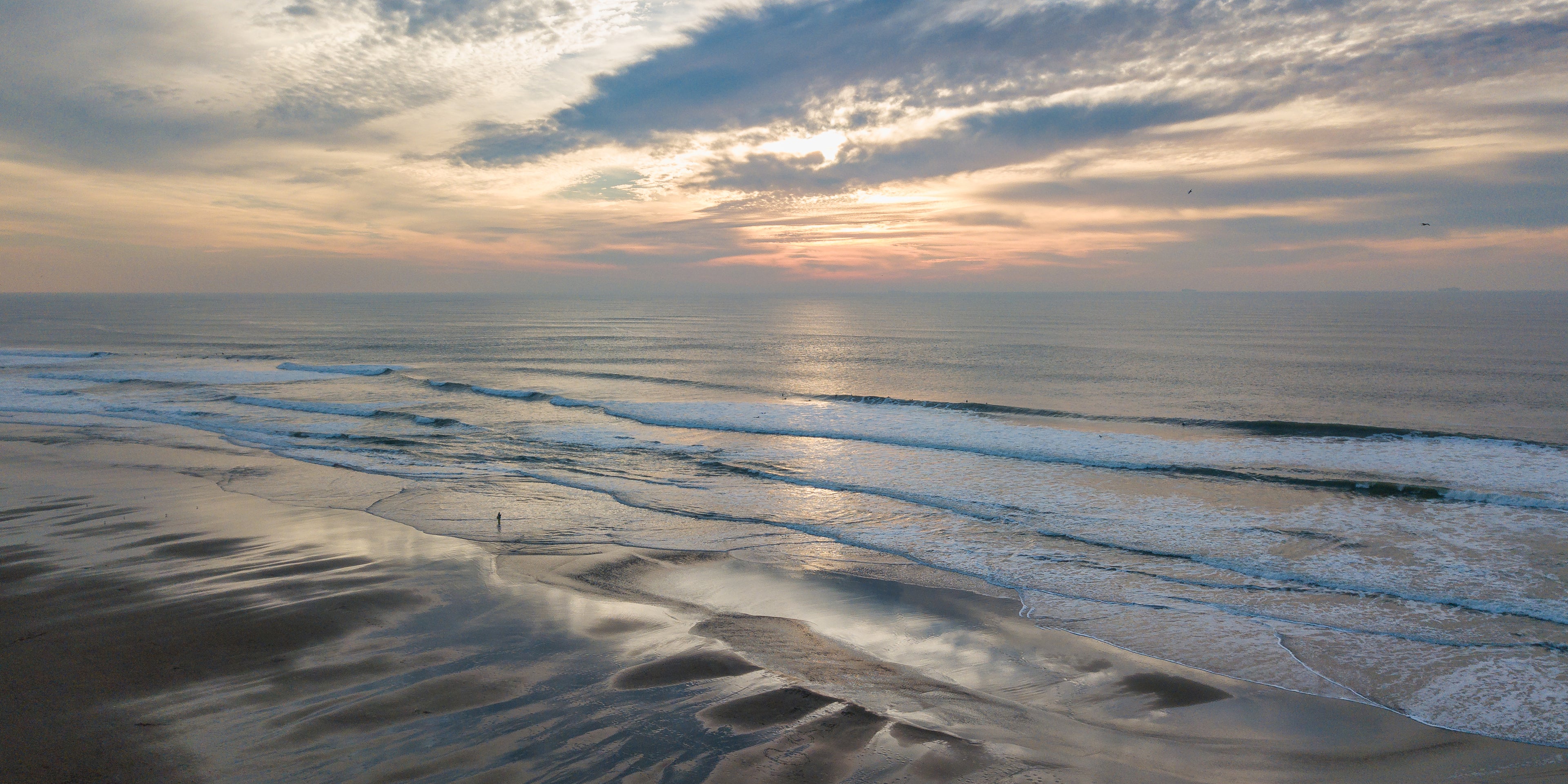 Aerial panoramic view of surfers entering the Pacific beneath a luminous sunset at Ocean Beach in San Francisco.