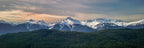Aerial panoramic photograph of snow-covered mountains rising above evergreen forest along British Columbia’s Sea-to-Sky Highway beneath a softly colored sky.