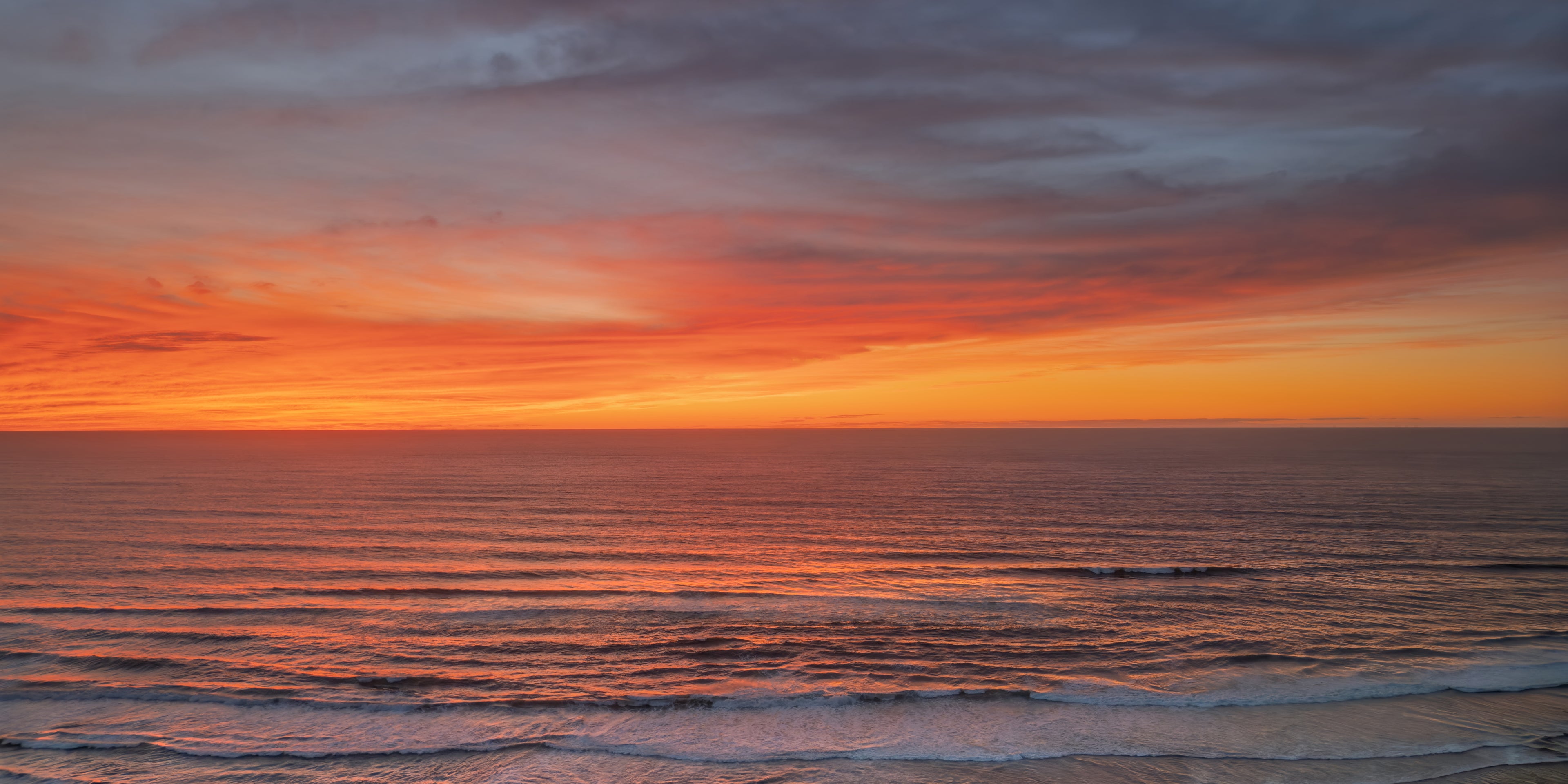 Brilliant orange and coral sunset clouds reflected across rhythmic ocean waves approaching the shore.