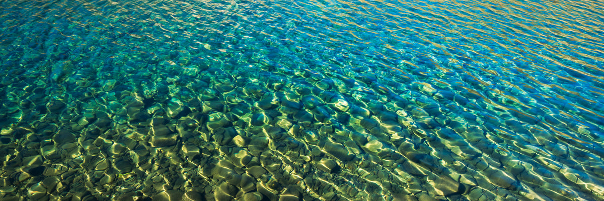 The Dance Below, a panoramic photograph of clear turquoise water with golden sunlight forming shifting patterns across submerged stones and surface ripples.