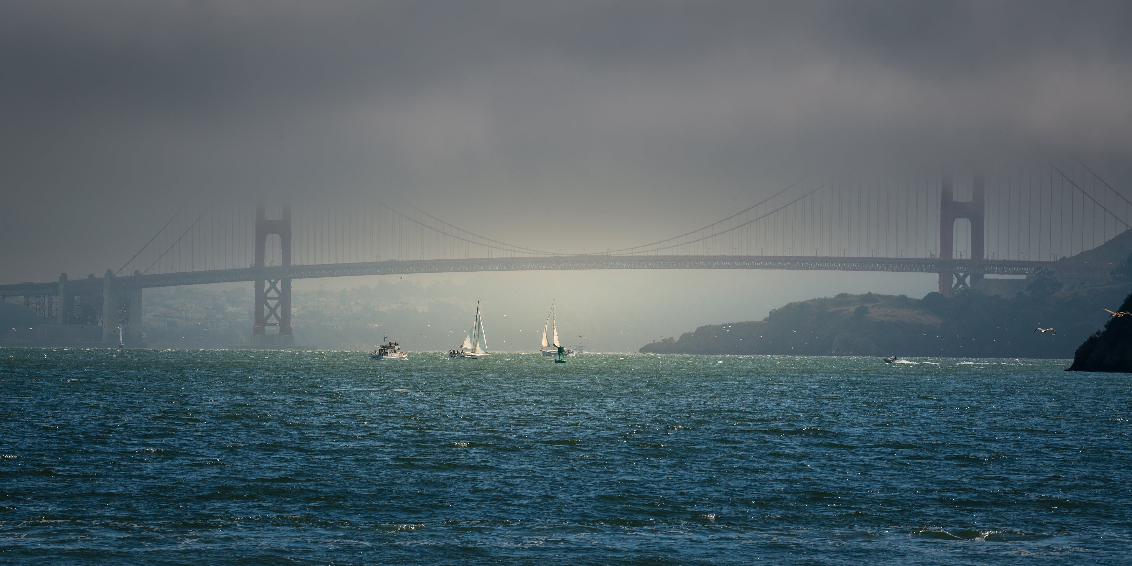 Sailboats pass through a break in the foggy light beneath the Golden Gate Bridge on San Francisco Bay.