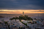 Coit Tower rising above San Francisco neighborhoods with the illuminated Bay Bridge beneath a glowing dawn sky.