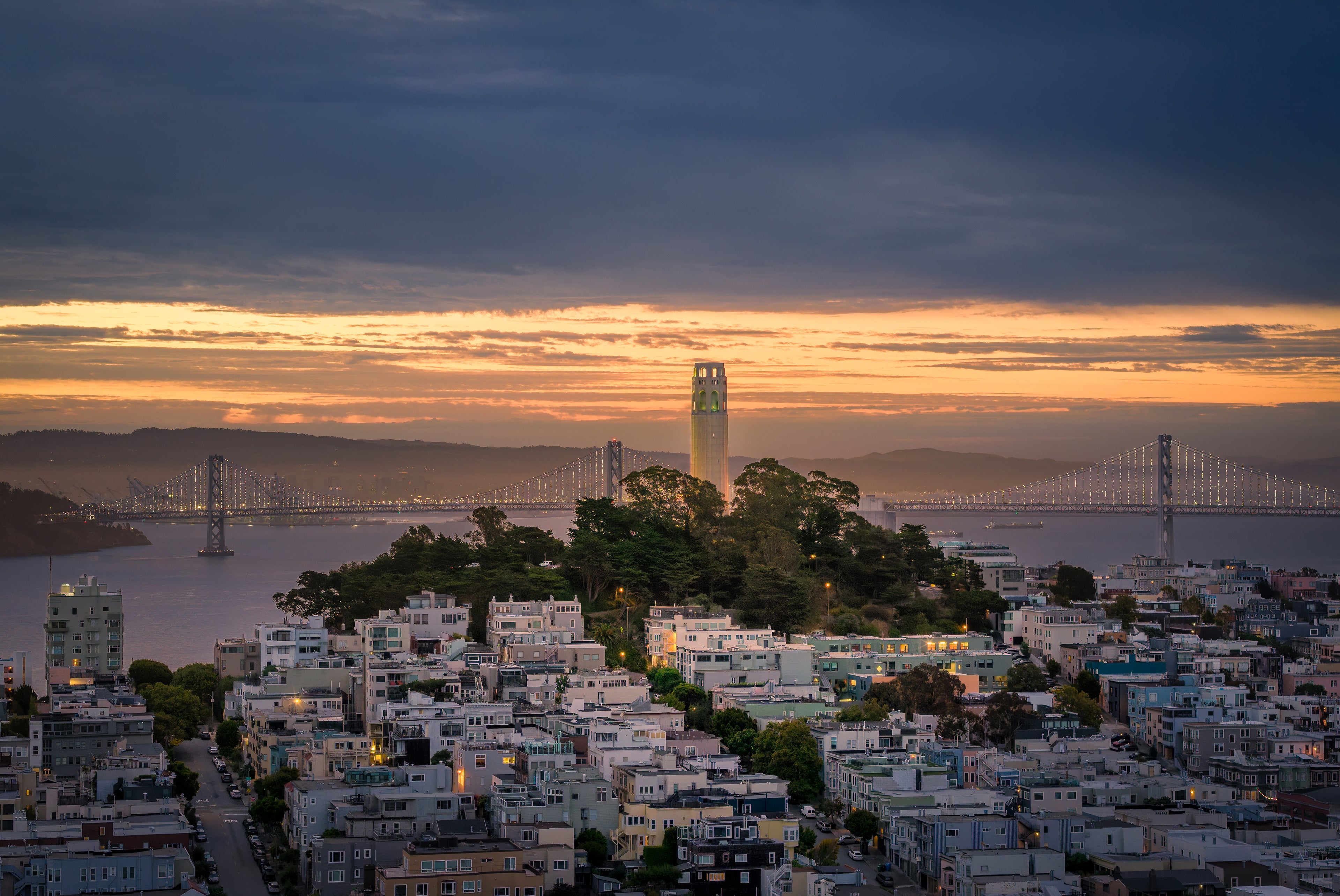 Coit Tower rising above San Francisco neighborhoods with the illuminated Bay Bridge beneath a glowing dawn sky.