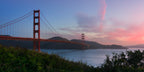 Golden Gate Bridge at dawn beneath a luminous sky of soft blue and rose, with calm water and shadowed foliage in the foreground.