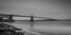 Black-and-white panoramic photograph of the Golden Gate Bridge and Fort Point emerging through soft morning fog above smooth water and a rocky shoreline.