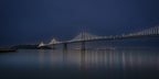 Panoramic early-morning photograph of the illuminated Bay Bridge emerging through light fog, with muted gold, blue, green, and rose reflections stretching across the still bay.
