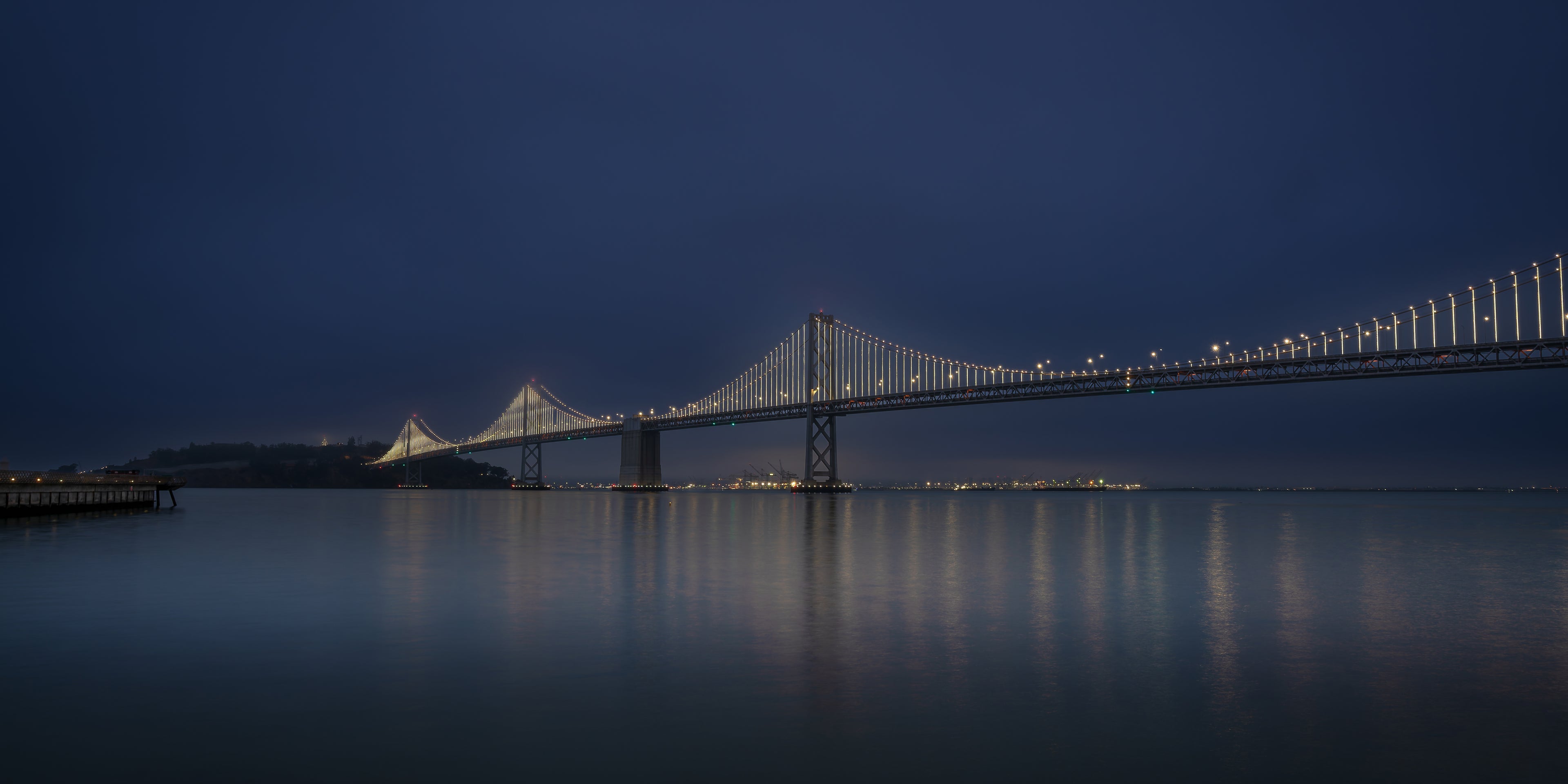 Panoramic early-morning photograph of the illuminated Bay Bridge emerging through light fog, with muted gold, blue, green, and rose reflections stretching across the still bay.
